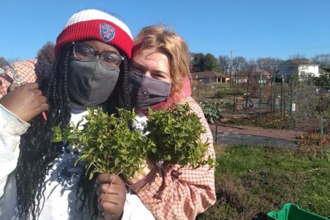 Two masked students embrace in a hug, holding greens.