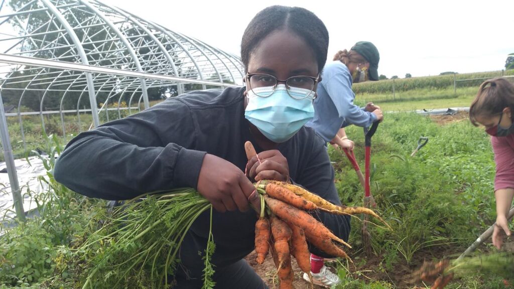 Teni Bakare ‘23, masked, holds a bunch of carrots.